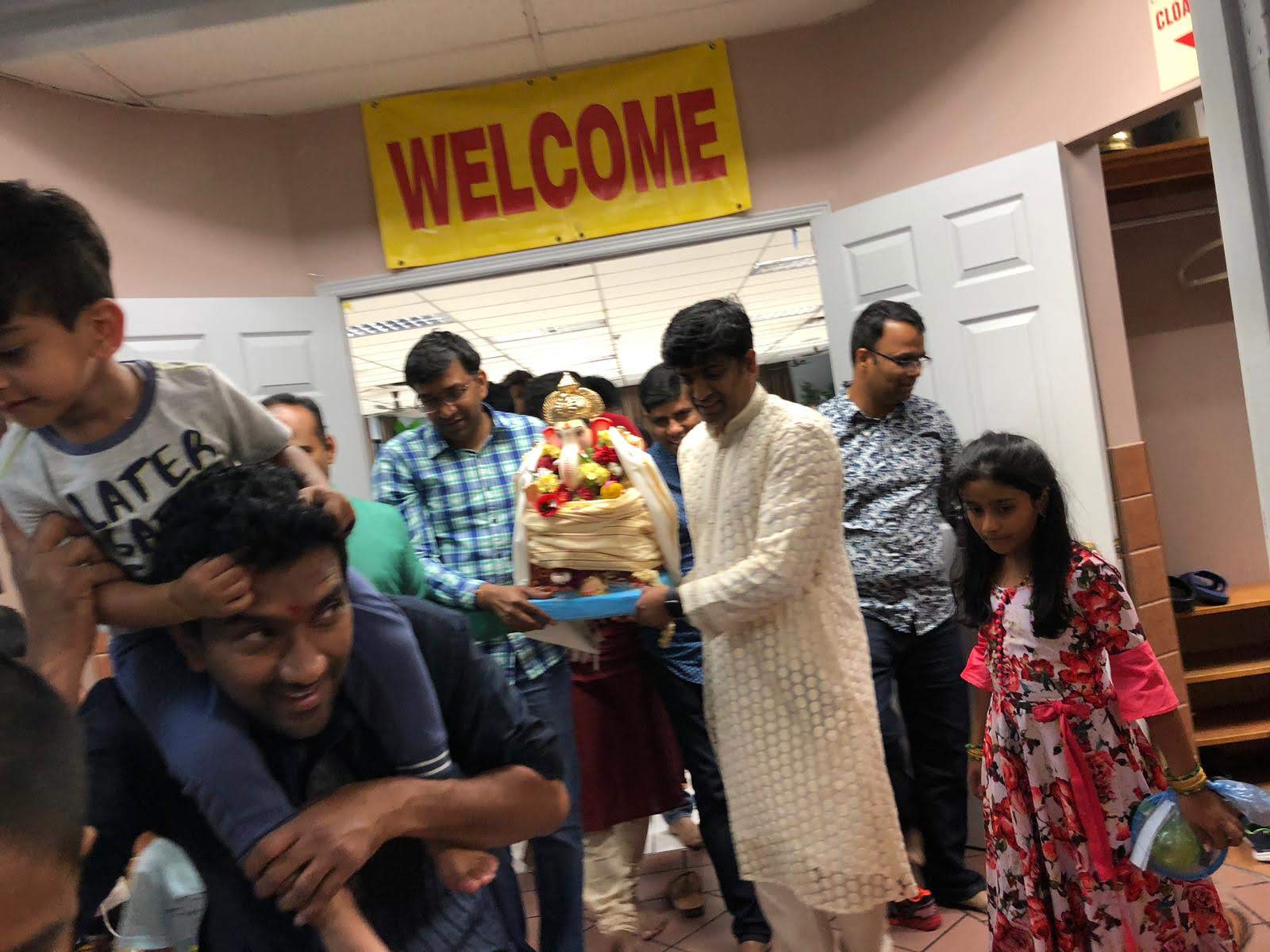 Community members carrying the Ganesh idol beneath a Welcome sign, 2019